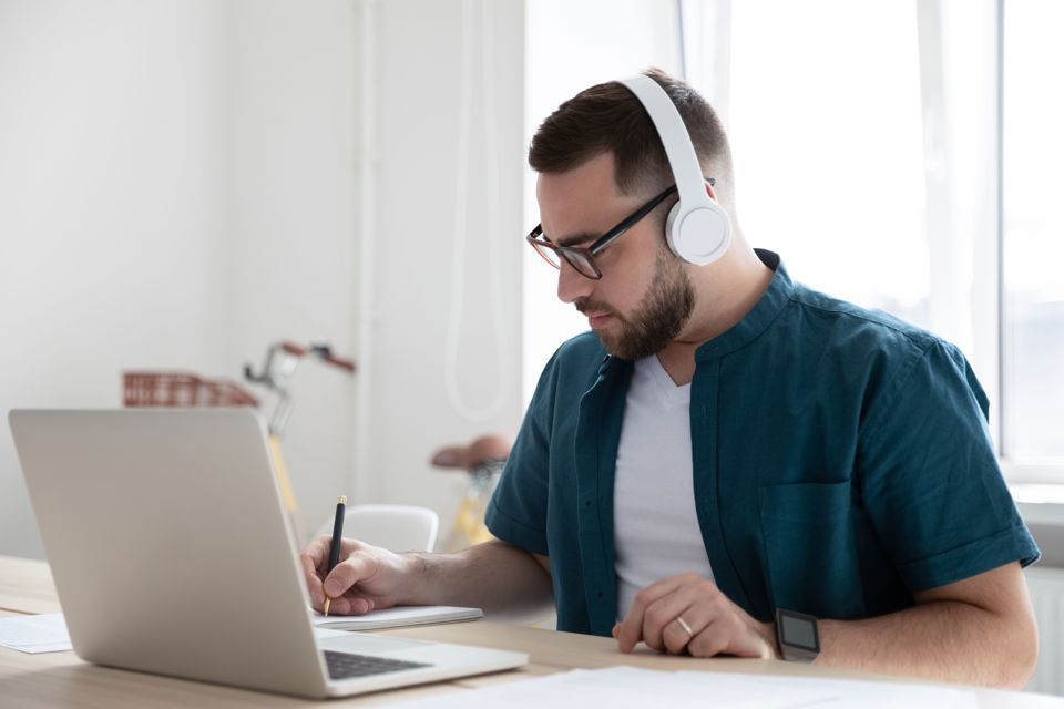 A male wearing headphones sitting at a desk and making notes.